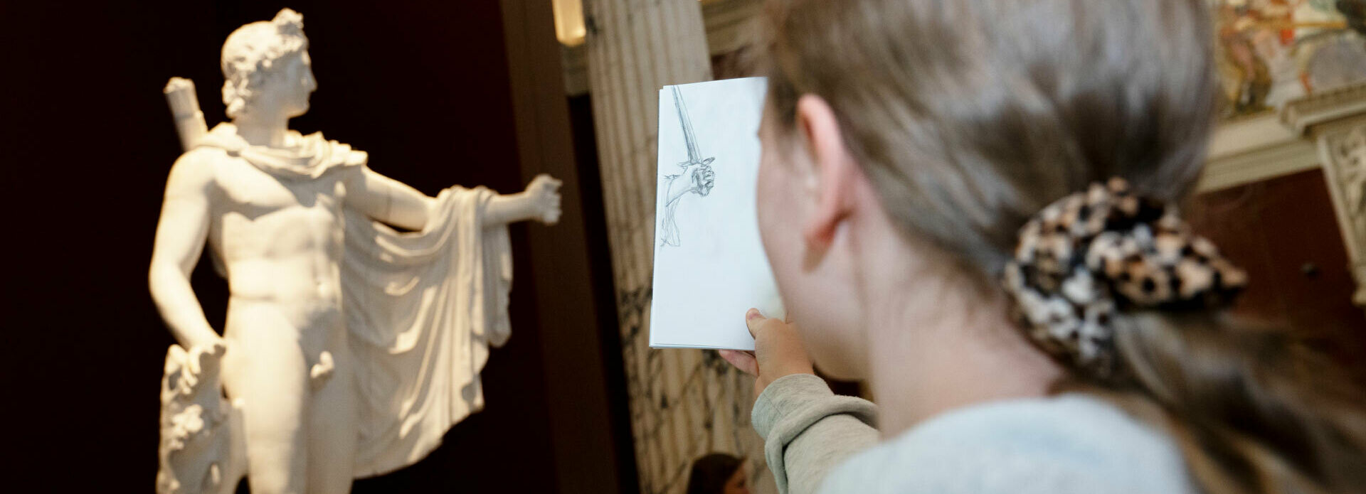 En flicka står framför en skulptur med ryggen mot betraktaren. Hon håller upp ett papper, där hon har ritat av en detalj av skulpturen, och jämför med förlagan.