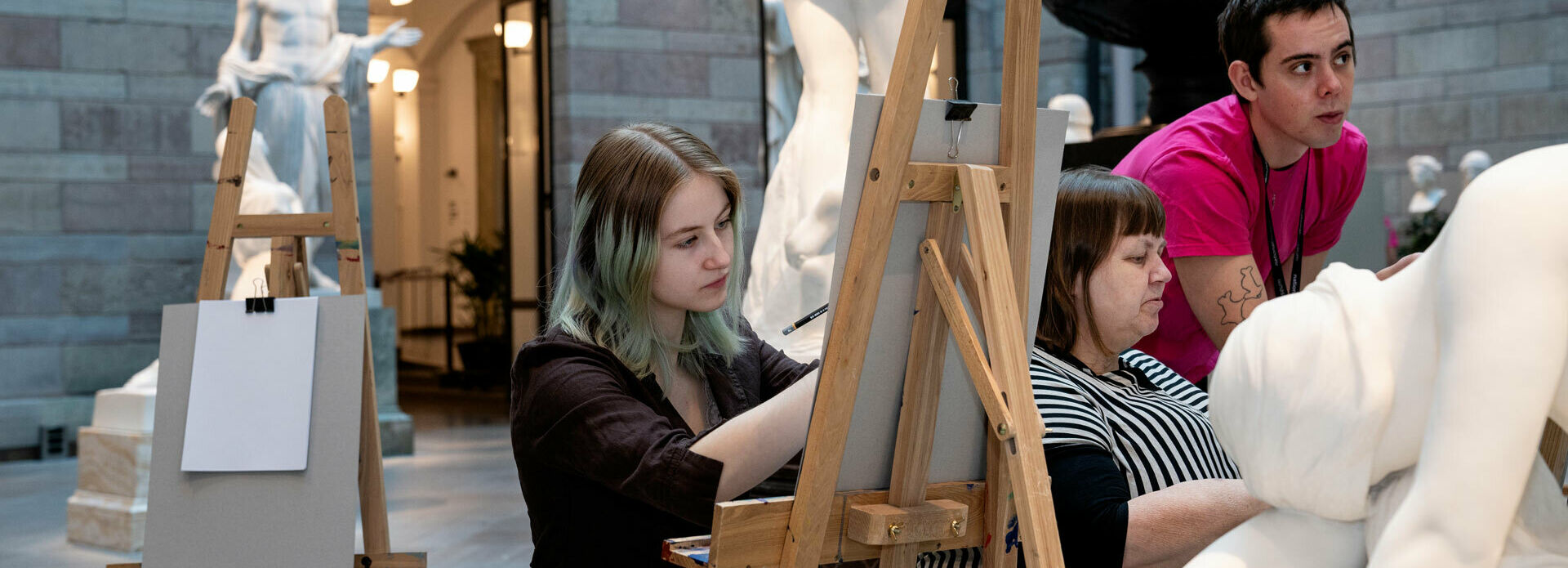 Två personer som siter vid stafflier och tecknar av skulpturer i en skulpturgård. En person står bredvid och ger råd.