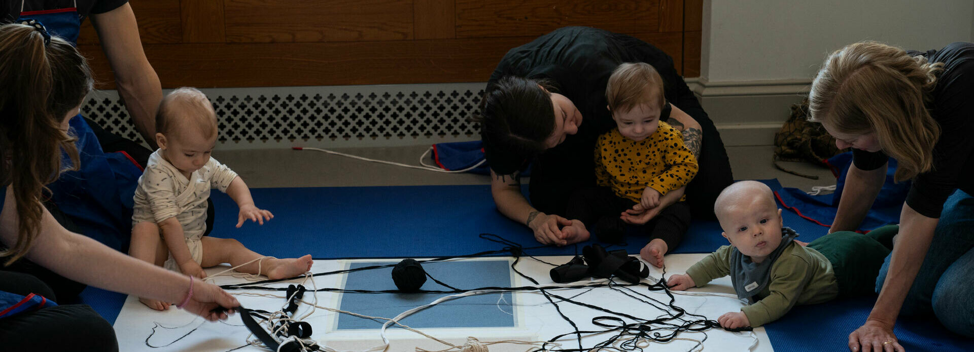 Småbarn med vuxna som kryper omkring på golvet. Där finns stora papper samt garnnystan och pennor som bebisarna ritar" med.