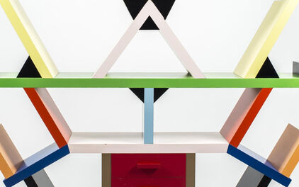 Geometrisk hylla i postmodern stil med färgglada former – rektanglar, trianglar och kvadrater i gult, blått, rött, grönt, svart och vitt – arrangerade asymmetriskt ovanpå en gråspräcklig bas. Designen är både funktionell och konstnärlig.