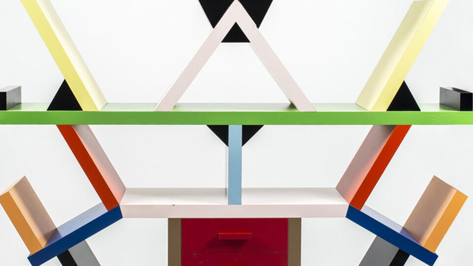Geometrisk hylla i postmodern stil med färgglada former – rektanglar, trianglar och kvadrater i gult, blått, rött, grönt, svart och vitt – arrangerade asymmetriskt ovanpå en gråspräcklig bas. Designen är både funktionell och konstnärlig.