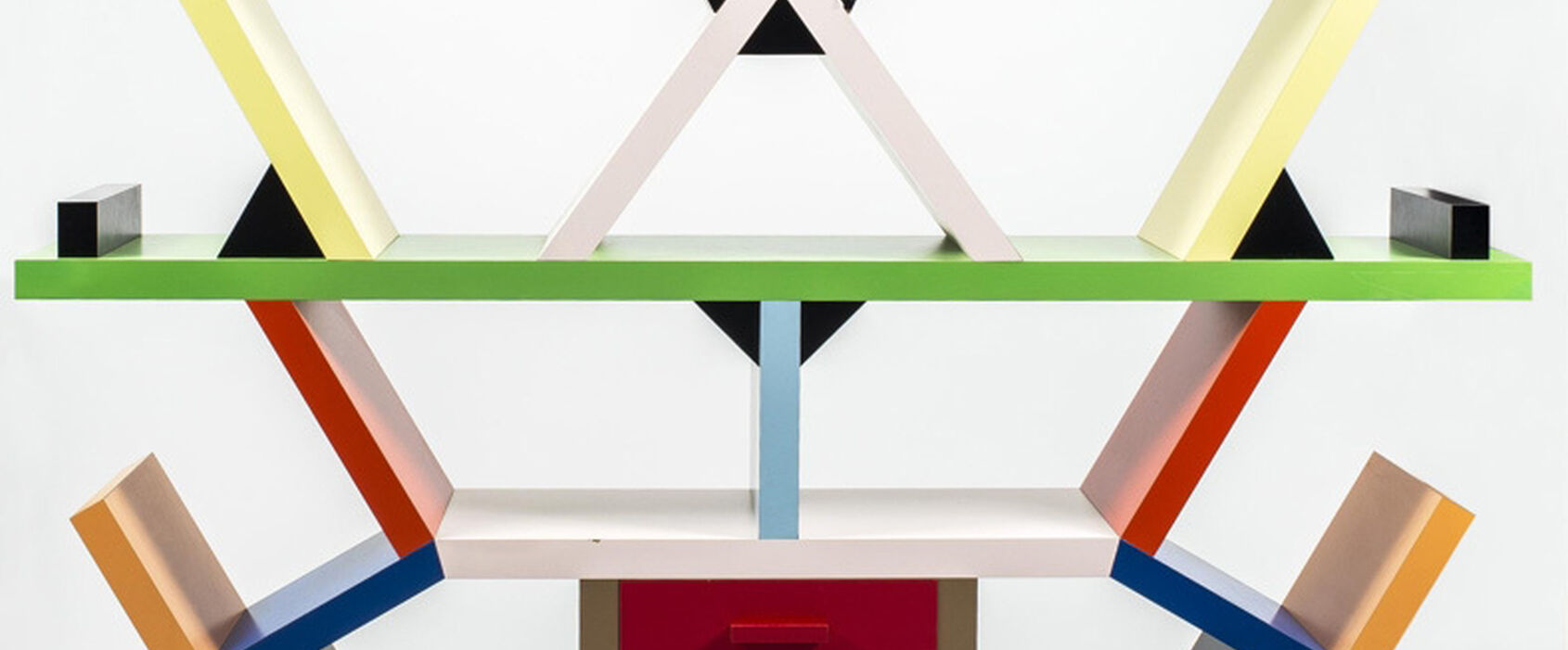 Geometrisk hylla i postmodern stil med färgglada former – rektanglar, trianglar och kvadrater i gult, blått, rött, grönt, svart och vitt – arrangerade asymmetriskt ovanpå en gråspräcklig bas. Designen är både funktionell och konstnärlig.
