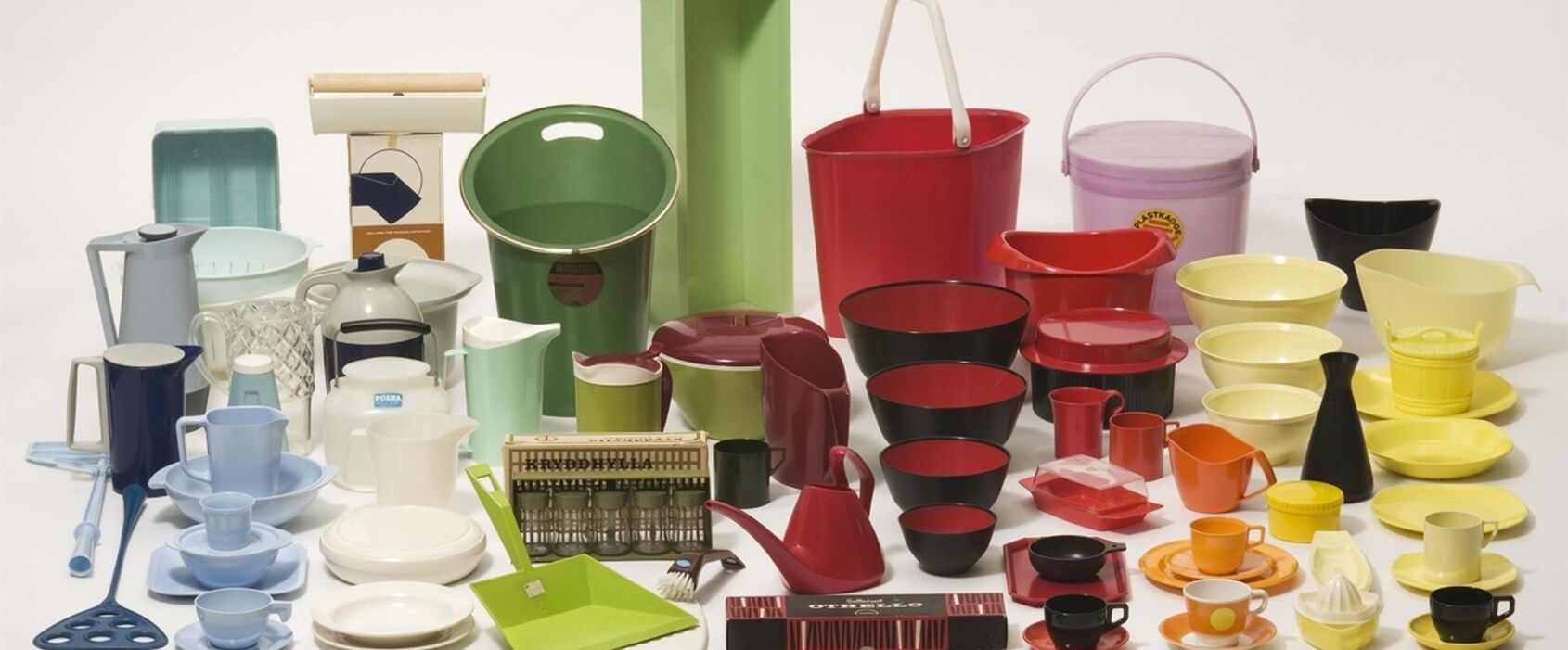 En samling färgglada plastföremål för hushållet – inklusive skålar, kannor, koppar, fat, brickor och vattenkannor – arrangerade tätt tillsammans på vit bakgrund. Föremålen är i retrostil och visar exempel på svensk plastdesign från 1900-talet.
