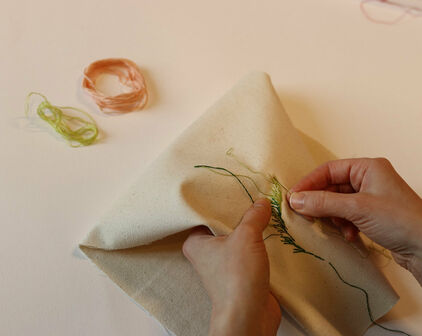 Händer som broderar ett blad med grön tråd på vitt tyg.