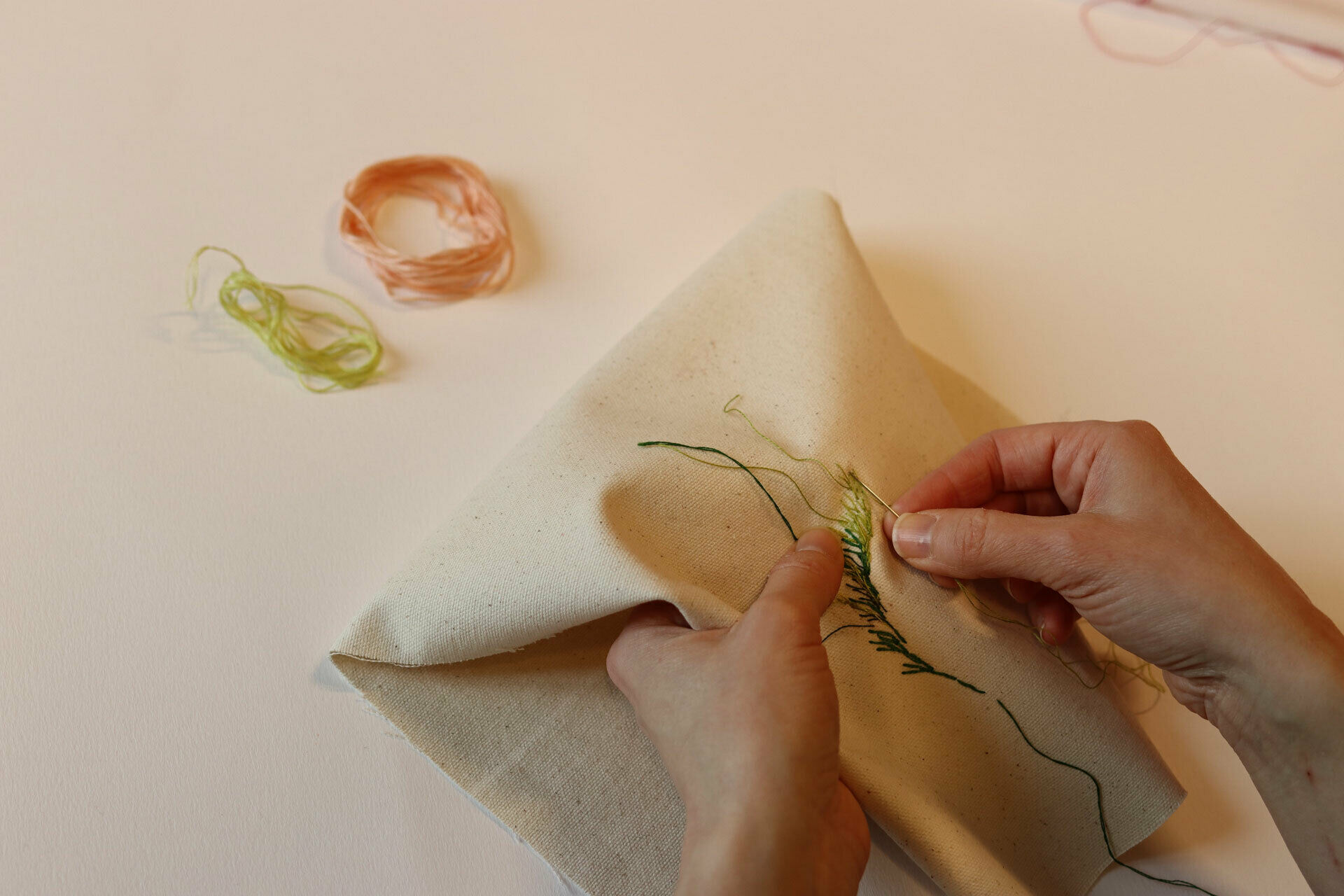 Händer som broderar ett blad med grön tråd på vitt tyg.
