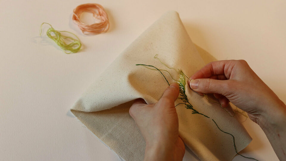 Händer som broderar ett blad med grön tråd på vitt tyg.