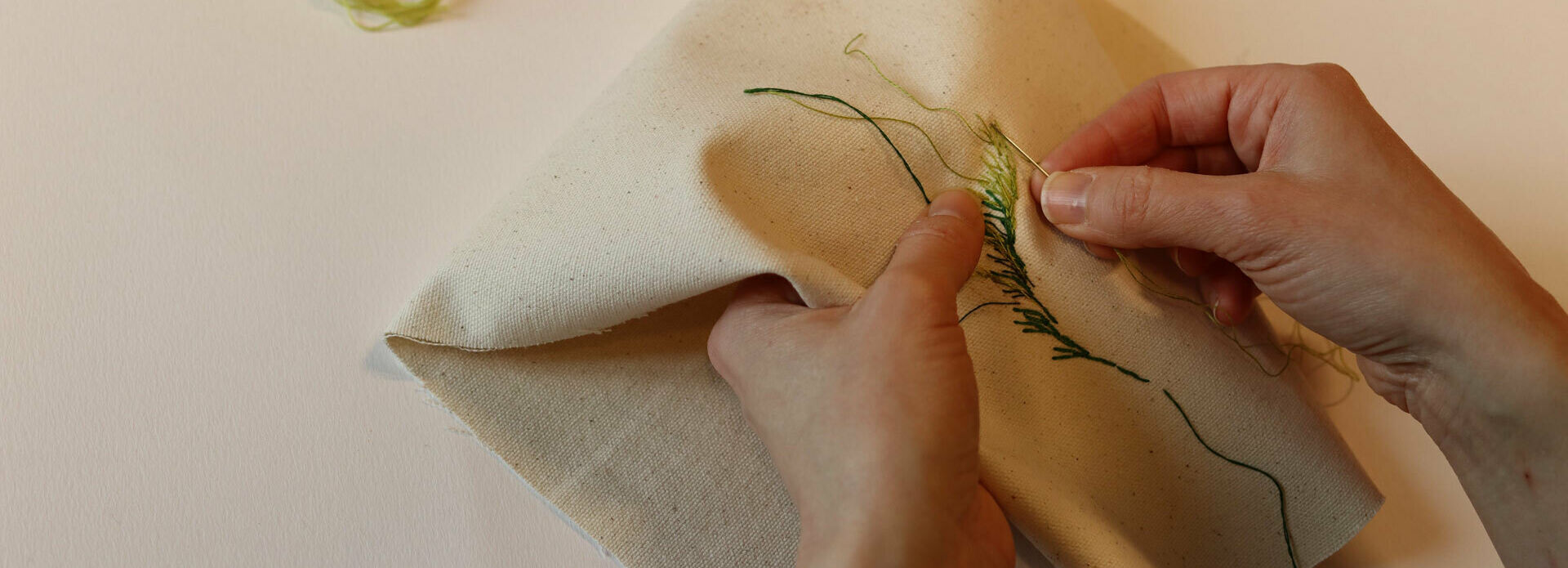 Händer som broderar ett blad med grön tråd på vitt tyg.