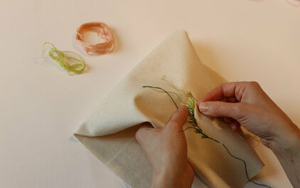 Händer som broderar ett blad med grön tråd på vitt tyg.