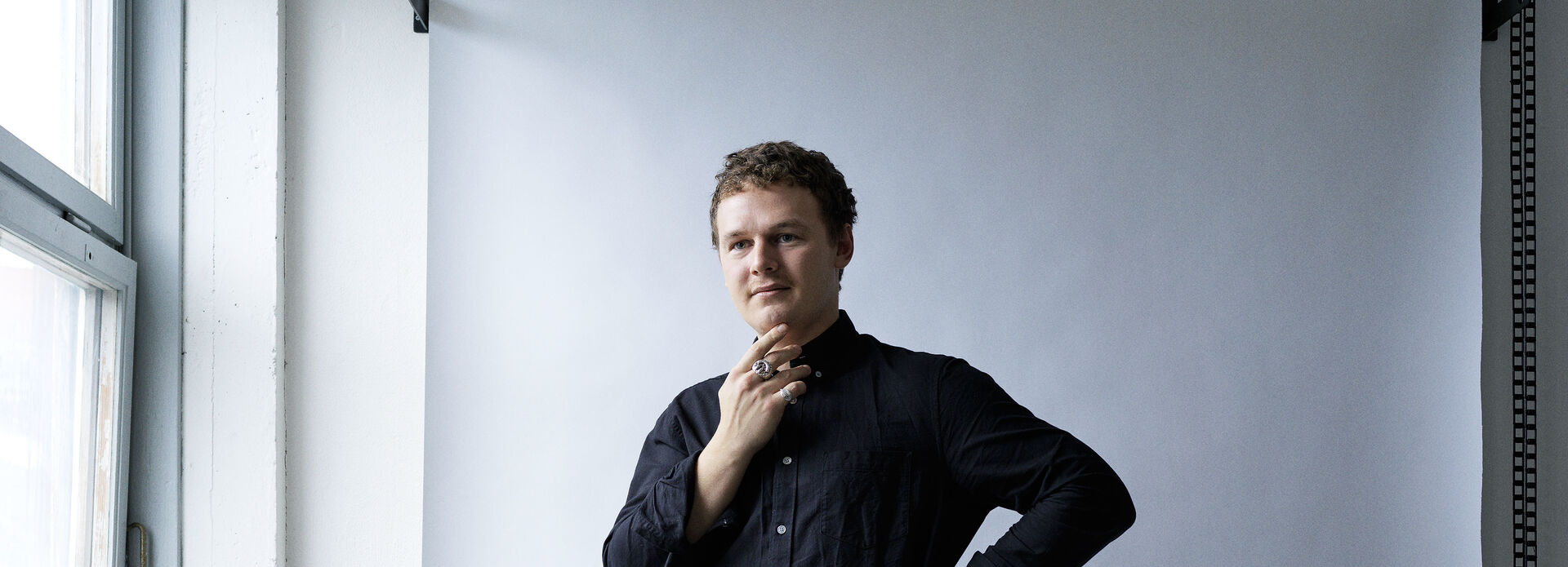 Porträtt av man med handen vid hakan. Mörka kläder, vit bakgrun.