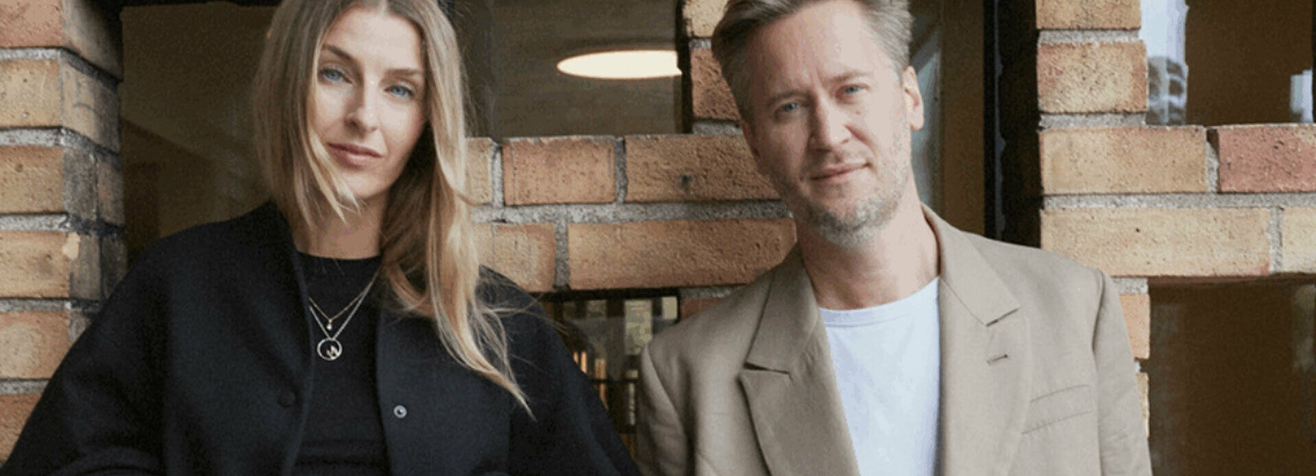 Fotografiskt dubbelporträtt av en kvinna och en man som står framför en tegelvägg.