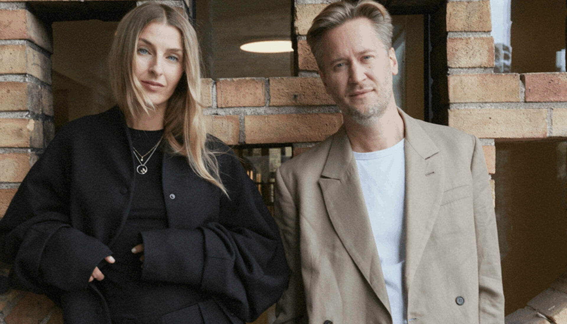 Fotografiskt dubbelporträtt av en kvinna och en man som står framför en tegelvägg.