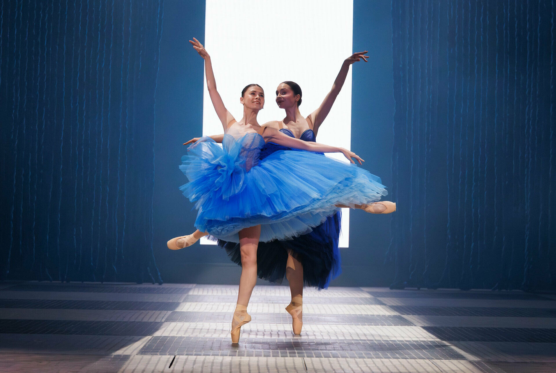 Foto av två balettdansöser, i Kungliga Operans balett Gustavia, som står på tåspetsen, iklädda klarblå tutu-klänningar.