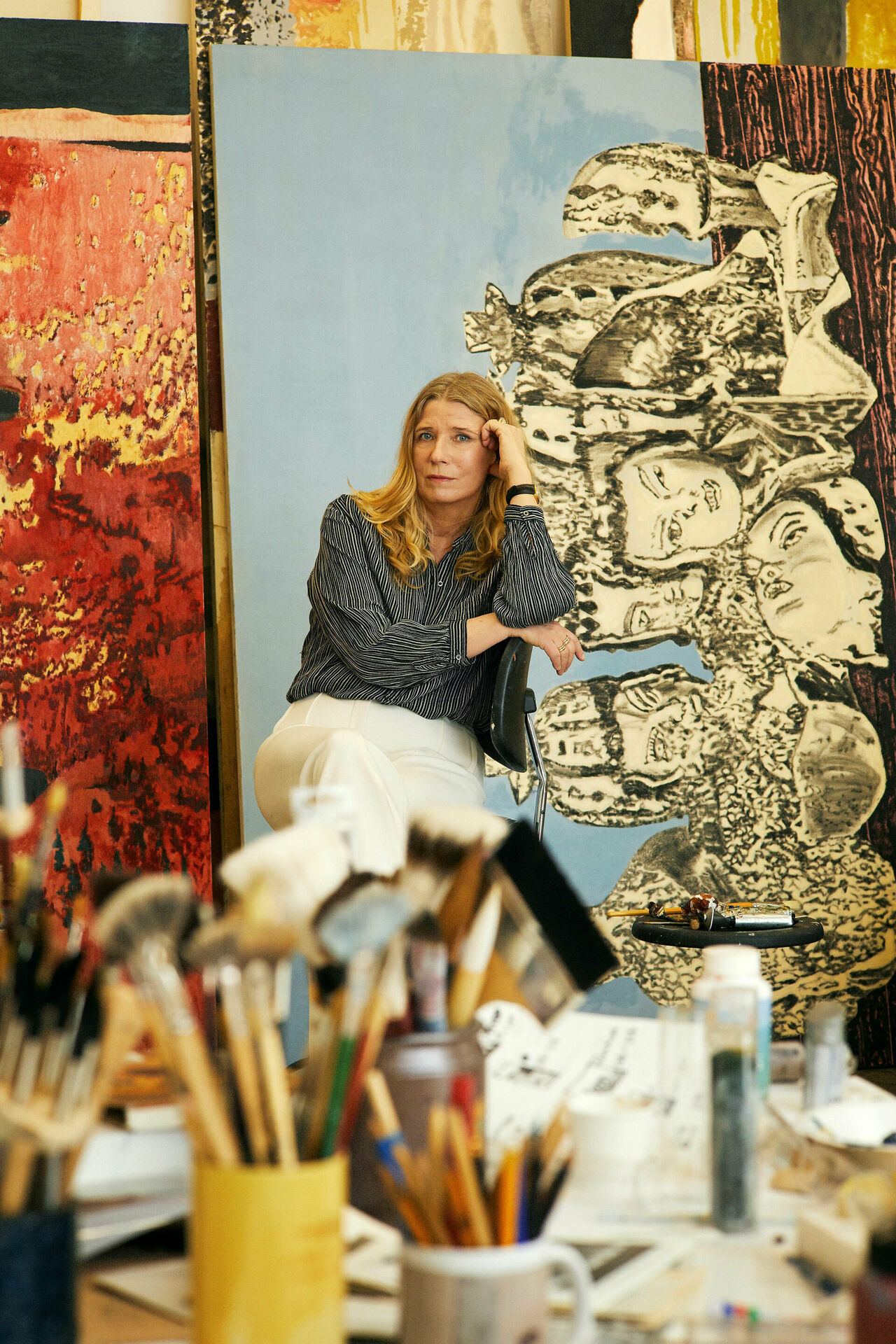 Fotografiskt porträtt av en kvinna - konstnären Karin Mamma Andersson - som sitter bland målningar i en ateljé