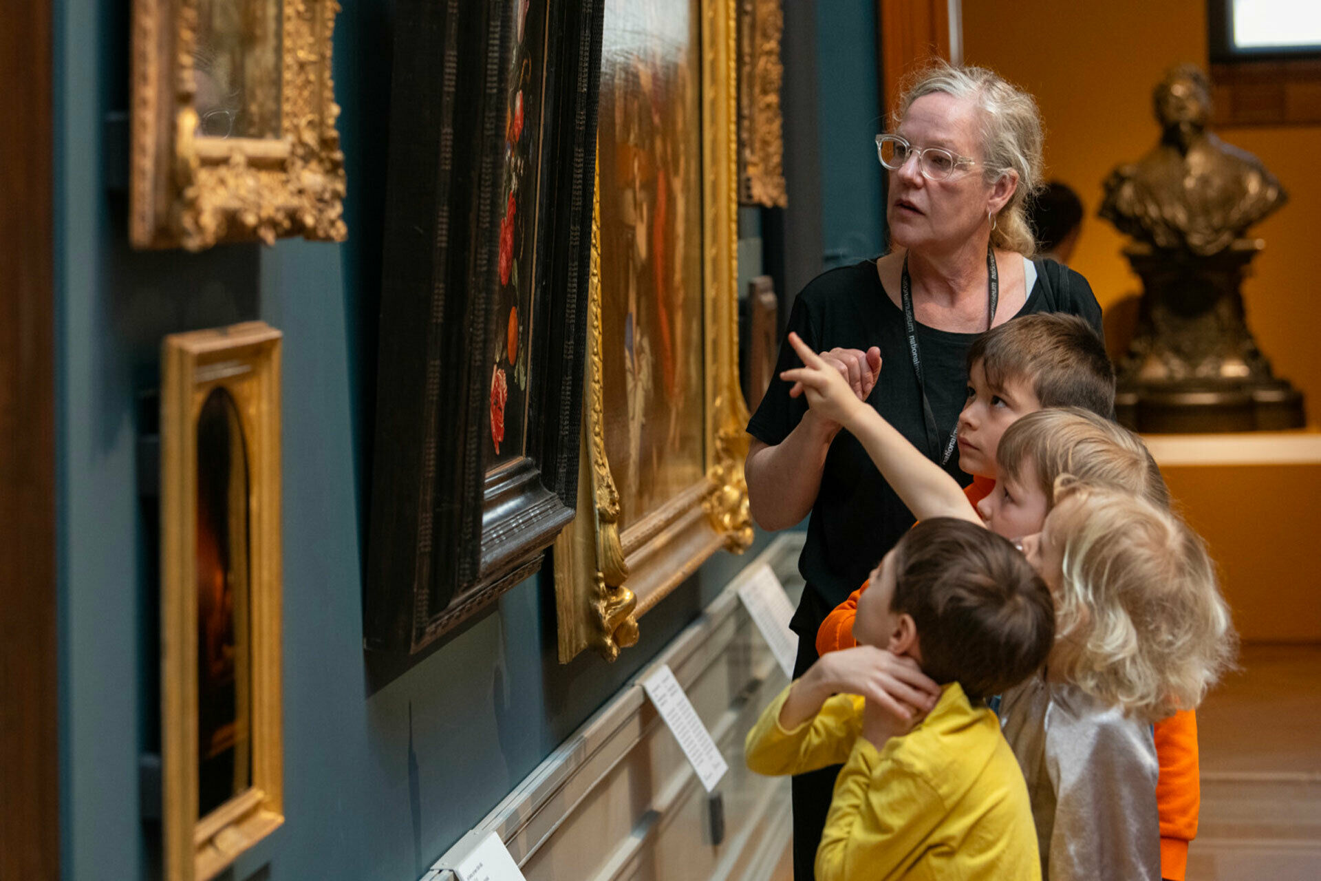 Några barn och en vuxen kvinna som tillsammans tittar på och pratar om tavlor på en vägg.