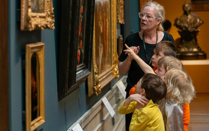 Några barn och en vuxen kvinna som tillsammans tittar på och pratar om tavlor på en vägg.