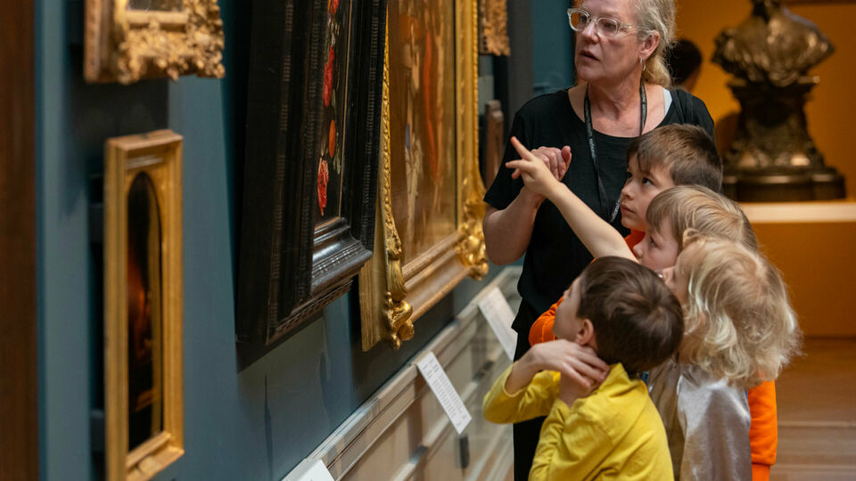 Några barn och en vuxen kvinna som tillsammans tittar på och pratar om tavlor på en vägg.