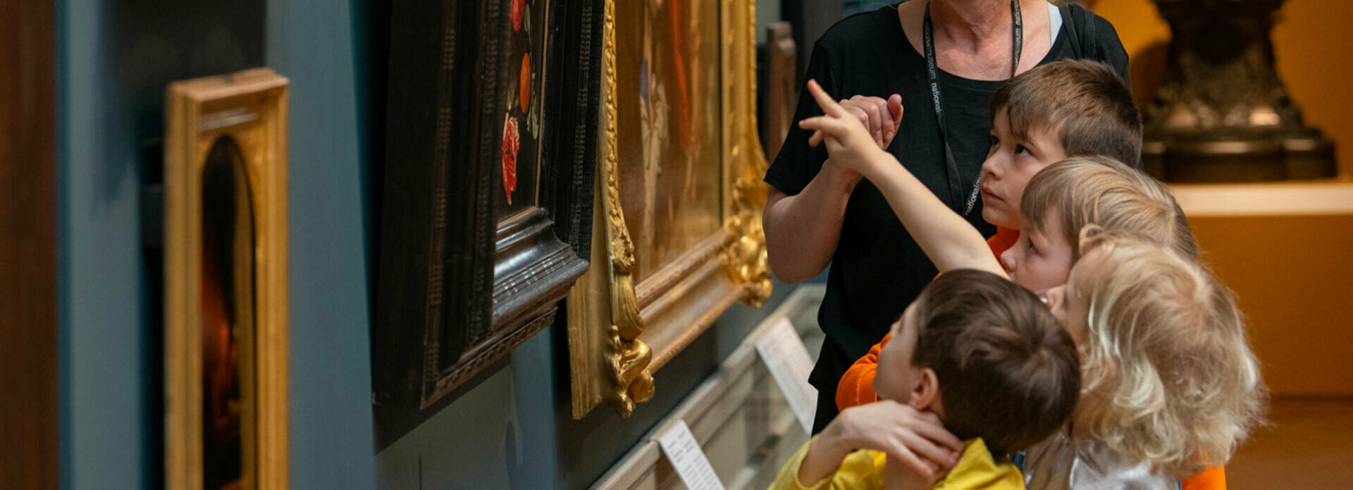 Några barn och en vuxen kvinna som tillsammans tittar på och pratar om tavlor på en vägg.