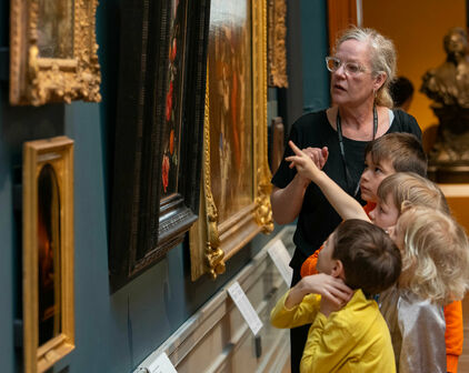 Några barn och en vuxen kvinna som tillsammans tittar på och pratar om tavlor på en vägg.